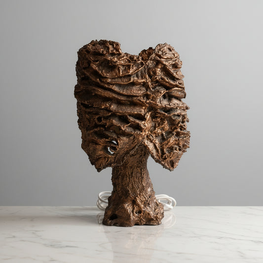 A sculptural wall-mounted lamp with a highly textured, organic brown and bronze metallic surface, shown mounted on a light-colored marble-patterned wall with a white cord visible.