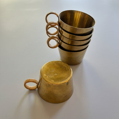 A set of brass cups with woven rattan handles, with one cup standing open in a stack and another cup lying upside down in the foreground on a light wood surface.