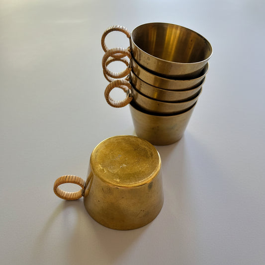 A set of brass cups with woven rattan handles, with one cup standing open in a stack and another cup lying upside down in the foreground on a light wood surface.